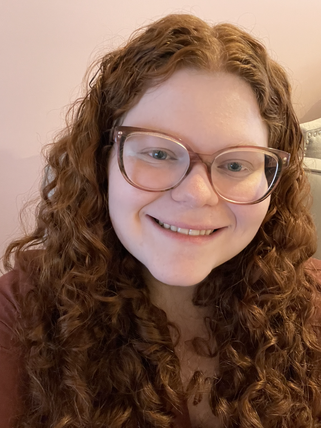 Picture of Laura. She is a white woman with curly red hair and big pink glasses. She is in front of a light pink background and is wearing a solid marroon shirt.