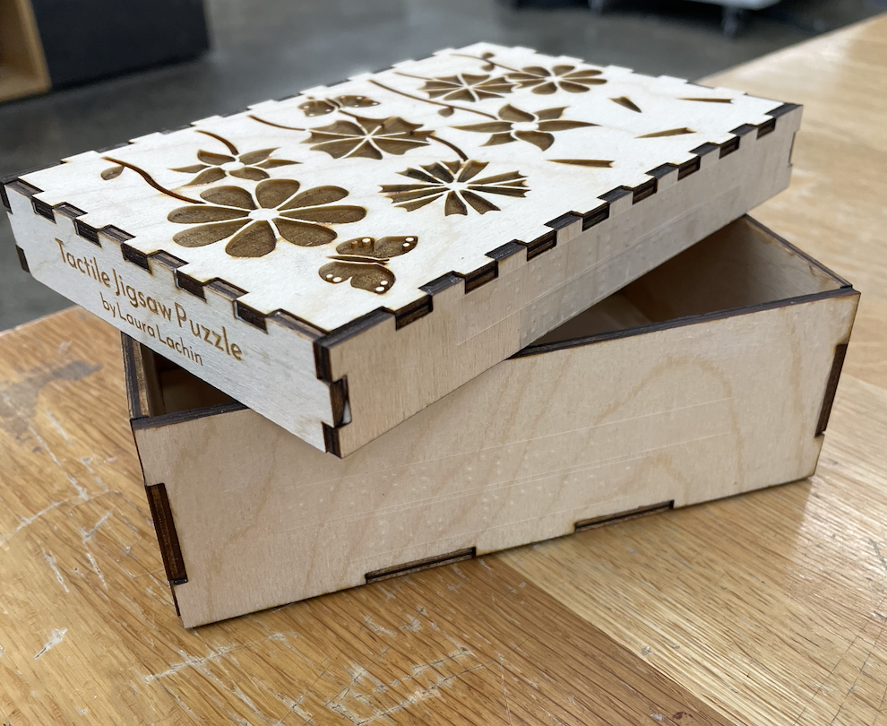 Three-fourths view of box with the lid resting diagonally on top of the bottom. Three sides of the lid are visible: the etched lid, one side that says 'Tactile Jigsaw Puzzle by Laura Lachin', and one side that has two Braille labels.