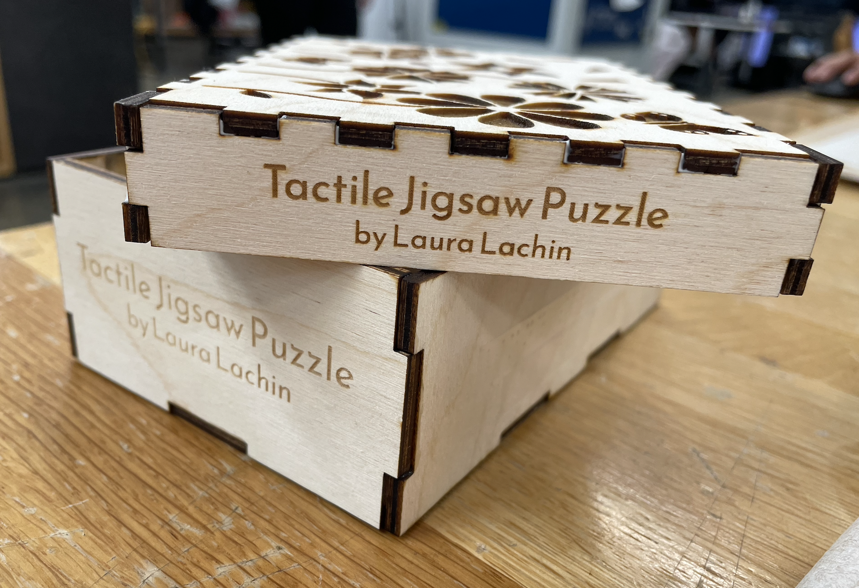 Side view of box with the lid resting diagonally on top of the bottom. The closest sides of both the box lid and bottom read 'Tactile Jigsaw Puzzle by Laura Lachin'.