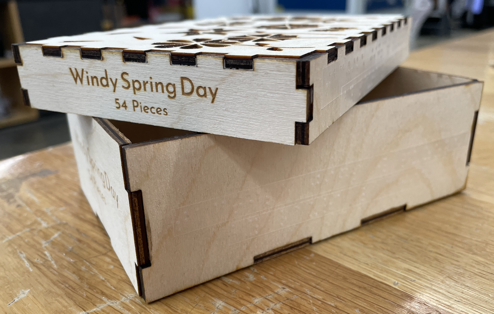 Three-fourths view of box with the lid resting diagonally on top of the bottom. In view are the box lid side that reads 'Windy Spring Day 54 Pieces' and one of the box bottom sides with Braille.